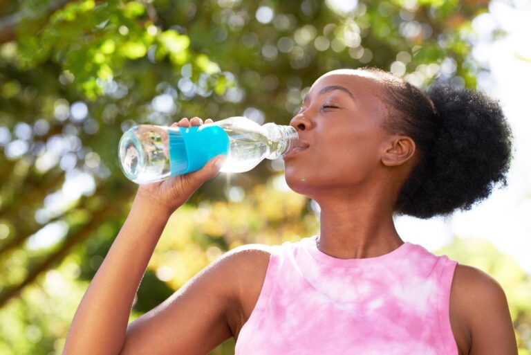 woman with her hair in a high ponytail wearing a pink and white blouse and holding a bottle of water to her lips